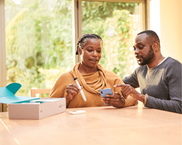 A middle aged man and women sit in conversation at their kitchen table. The woman has the Tempo Pen and Tempo Smart Button in her right hand, the man is pointing at the screen of the smartphone in her left hand